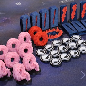 Op de afbeelding: Een verzameling spelonderdelen in verschillende vormen en kleuren. Er zijn roze, donutvormige stukken, blauwe rechthoekige stukken met rode accenten, rode cirkelvormige stukken en zilveren en witte cirkelvormige stukken. De stukken zijn gerangschikt op een donkerblauwe ondergrond.