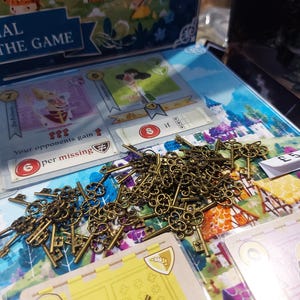 以下が含まれることがあります： ボードゲーム。カードと、アンティーク風のブロンズ色の鍵の山。背景には「AL THE GAME」の文字が入ったゲームボックスが見える。カードにはイラストとテキストがあり、「6」の数字と「if」の文字が見える。