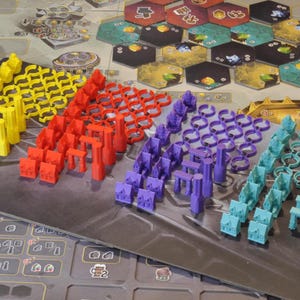 May include: A board game setup with various colorful game pieces. Yellow, red, purple, and teal pieces are arranged in rows. The game board features hexagonal tiles and detailed artwork, suggesting a strategy or resource management theme.