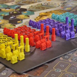 May include: A board game setup with various colorful game pieces. The pieces include yellow, red, purple, and teal miniature buildings and towers. The game board features hexagonal tiles and a dark gray playing surface.
