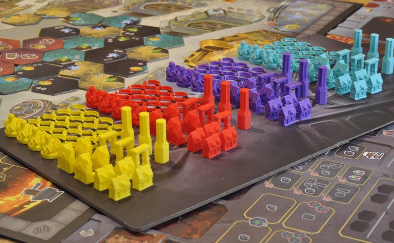 May include: A board game setup with various colorful game pieces. The pieces include yellow, red, purple, and teal miniature buildings and towers. The game board features hexagonal tiles and a dark gray playing surface.