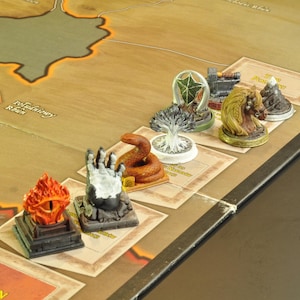 Puede incluir: Un juego de mesa con varias figuras en miniatura. Las figuras incluyen un ojo llameante, una mano, una serpiente, un árbol, un edificio y una montaña. El tablero del juego tiene un diseño de mapa con el texto "Refuging Ranch" y "Stan Wo".