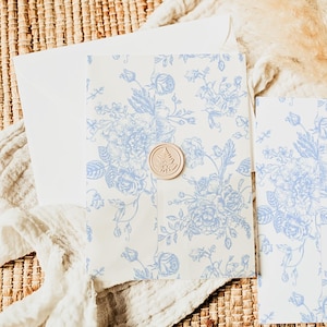 May include: An invitation suite featuring a white envelope and stationery with a blue floral pattern. A round, light brown wax seal with a tree design secures the invitation. The stationery is arranged on a woven surface.