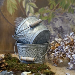 Könnte beinhalten: Zwei graue Metall-Eimer mit dekorativen Mustern und Holzgriffen. Die Eimer sind übereinander gestapelt und stehen auf einem Moosbett. Im Hintergrund befinden sich getrocknete Blumen.