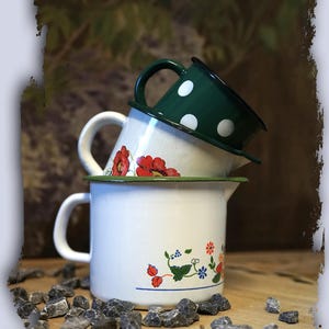 Könnte beinhalten: Drei Emaille-Becher, die übereinander gestapelt sind. Der obere Becher ist grün mit weißen Punkten, der mittlere Becher ist weiß mit roten Mohnblumen und der untere Becher ist weiß mit einem grünen, blauen und roten Blumendesign.