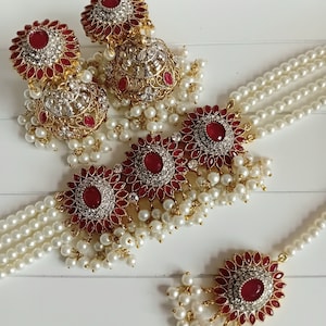 May include: A traditional Indian jewellery set featuring a gold-toned choker necklace with red and white stones, a matching pair of earrings, and a small pendant. The necklace and earrings are adorned with pearl accents.