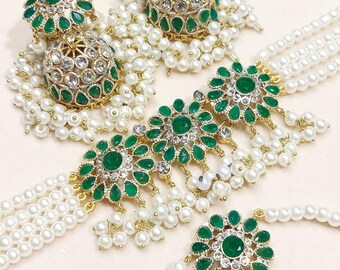 May include: A set of green and gold jewellery with pearl accents. The set includes a necklace, earrings, and a maang tikka. The necklace is a choker style with a green and gold pendant in the centre. The earrings are dangle earrings with a green and gold pendant. The maang tikka is a small, green and gold pendant that is worn on the forehead.