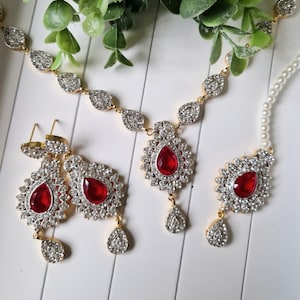 May include: A gold-coloured jewellery set comprising a necklace, earrings, and a headpiece. Each item features a central red, teardrop-shaped stone encircled by clear, sparkling crystals. The necklace and earrings have a floral design.