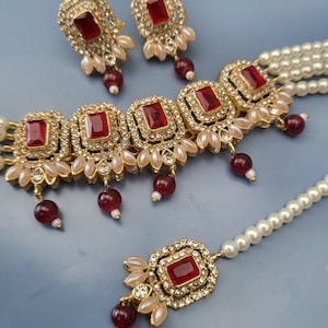May include: A gold-coloured jewellery set comprising a necklace, earrings, and a headpiece. The set features square red gemstones, surrounded by clear crystals and small pearls. The necklace and headpiece have dangling red beads.