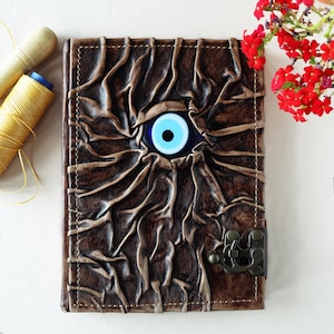 Peut inclure: Un journal en cuir marron avec un motif d'œil décoratif et un fermoir en métal. L'œil est bleu et blanc avec une pupille noire.