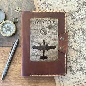 Könnte beinhalten: Ein braunes Lederjournal mit einem rechteckigen Einsatz, der das Wort AVIATOR, eine Windrose und die Silhouette eines Flugzeugs auf einem Kartenhintergrund zeigt. Das Journal hat einen Seitenriemen mit zwei Druckknöpfen und sichtbaren Nähten.