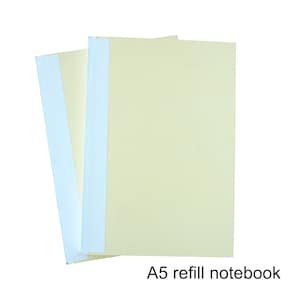Puede incluir: Una pila de cuadernos de recambio A5 con páginas de color amarillo pálido y lomos azul claro. Los cuadernos están apilados, con el superior ligeramente desplazado. El texto "A5 refill notebook" está en la parte inferior.