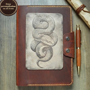 Op de afbeelding: Een bruin lederen dagboek met een rechthoekig inzetstuk met een gedetailleerde zwart-witillustratie van een opgerolde slang. Het dagboek heeft een zijriem vastgezet met twee metalen drukknopen, een houten pen en een opgerolde perkamenten rol.
