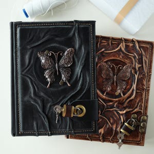 May include: Two leather journals with butterfly embellishments. One journal is black with a clasp closure and key. The other journal is brown with a lock and key. Both journals have embossed butterfly designs.