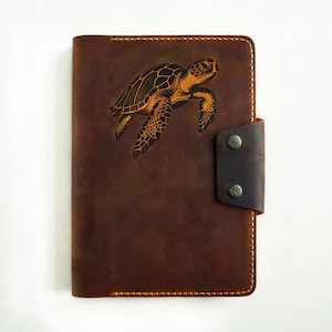 May include: A brown leather journal with a stitched border and a flap closure. The front cover features an engraved image of a sea turtle.
