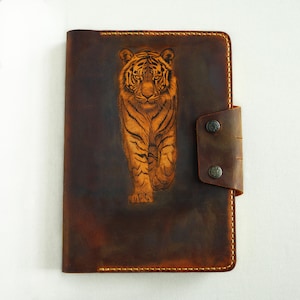 May include: A brown leather journal with a detailed orange tiger engraving on the front cover. The journal has a leather strap closure with two metal button studs. The edges are stitched with a light brown thread.