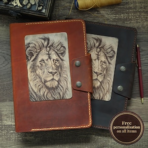 May include: Two leather journals, one tan and one dark brown, each featuring a rectangular inset panel with a detailed etched lion portrait. Both journals have a side flap secured by two metal snaps and are accompanied by a red pen.