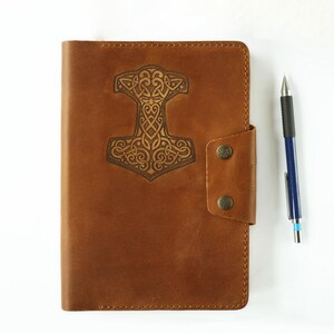 May include: A brown leather journal with a decorative embossed design of a hammer with a Celtic knot pattern. The journal has a flap closure with a snap button.