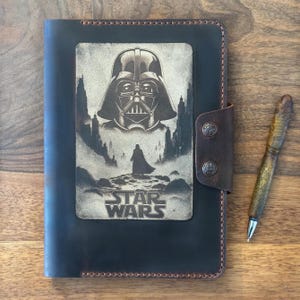 Könnte beinhalten: Dunkelbraunes Lederjournal mit einem Star Wars-Design. Das Journal zeigt ein geprägtes Bild von Darth Vader und dem Star Wars-Logo. Ein passender Stift mit braunem und goldenem Muster liegt neben dem Journal.