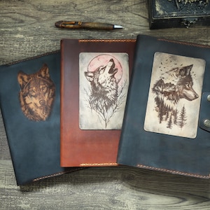 Könnte beinhalten: Drei Lederjournale mit Wolfskunst. Die Journale sind in Braun- und Blautönen gehalten und zeigen detaillierte Illustrationen von Wölfen und Naturszenen. Ein Stift liegt auf der Holzoberfläche.