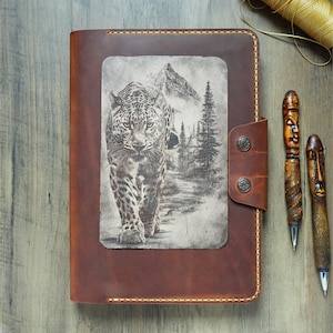 May include: A brown leather journal featuring a rectangular inset with a detailed black and white illustration of a leopard walking through a forest toward a mountain. The journal has a side flap secured by two metal snaps and visible stitching.