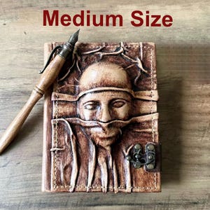 May include: A brown leather journal with a carved face design and a metal clasp. A wooden pen with a metal nib rests beside the journal. The text "Medium Size" is visible at the top of the image.