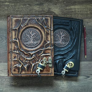 Könnte beinhalten: Zwei Lederjournale mit Lebensbaum-Designs. Eines ist braun mit Schloss und Schlüssel, das andere schwarz mit einem Verschluss. Ein bordeauxfarbener Stift liegt daneben auf einer Holzoberfläche.