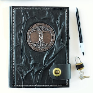May include: Black leather journal with a tree of life emblem, white stitching, and a clasp. A black pen and a small padlock with keys are next to the journal. The journal's cover has a textured, wrinkled appearance.