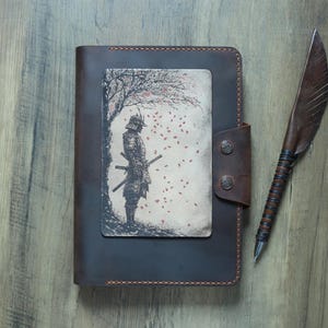 Può includere: Un diario in pelle marrone con un'illustrazione di samurai sulla copertina. Il diario ha una chiusura a bottone ed è abbinato a una penna. Il samurai è in piedi sotto un albero con foglie rosse che cadono.