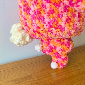 May include: A pink, orange, and white crocheted plush toy with a fluffy texture. The toy has a round body and three white pom-poms for feet.