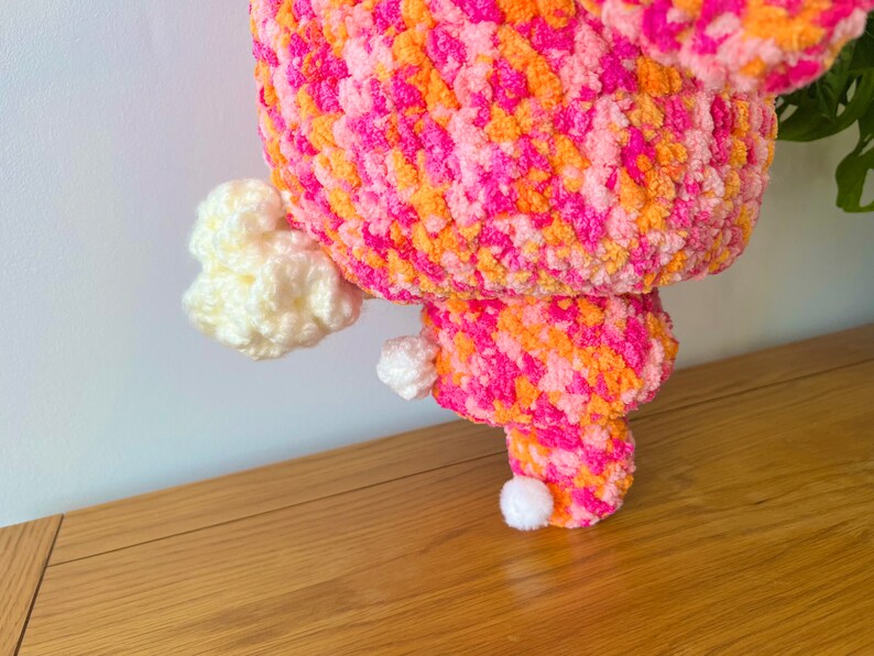 May include: A pink, orange, and white crocheted plush toy with a fluffy texture. The toy has a round body and three white pom-poms for feet.
