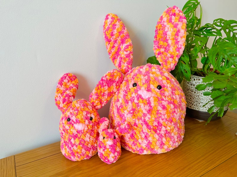 May include: Three crocheted bunnies in pink and orange. The largest bunny is sitting upright with its ears up. The middle bunny is sitting with its ears down. The smallest bunny is sitting with its ears down.