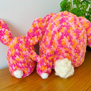 May include: Two pink and orange crocheted bunny plushies with white fluffy tails. The bunnies are lying on a wooden surface.