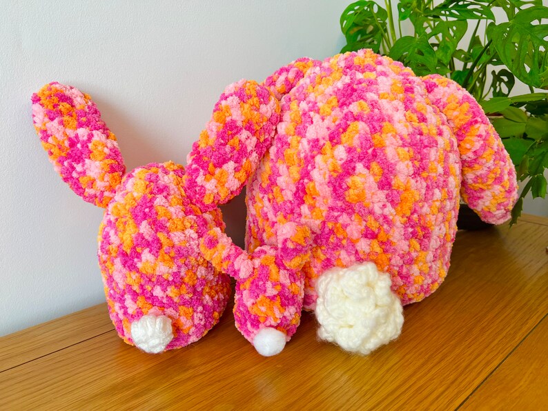 May include: Two pink and orange crocheted bunny plushies with white fluffy tails. The bunnies are lying on a wooden surface.