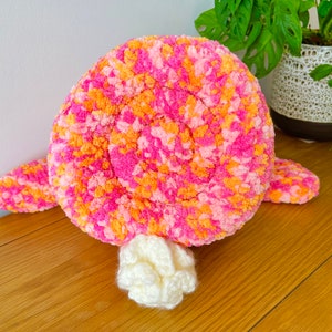 May include: A pink and orange crocheted plush toy shaped like a turtle. The turtle has a white crocheted tail.