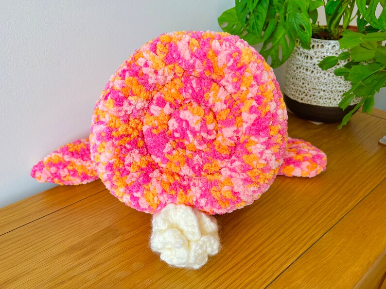 May include: A pink and orange crocheted plush toy shaped like a turtle. The turtle has a white crocheted tail.