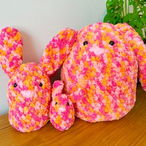 May include: Three crocheted bunny plushies in pink and orange. The largest bunny is sitting upright, while the two smaller bunnies are lying down.