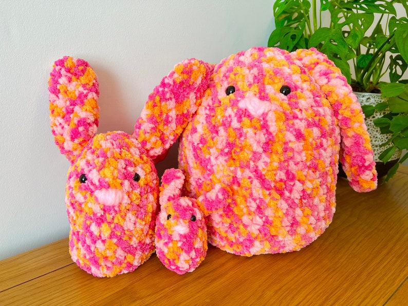 May include: Three crocheted bunny plushies in pink and orange. The largest bunny is sitting upright, while the two smaller bunnies are lying down.
