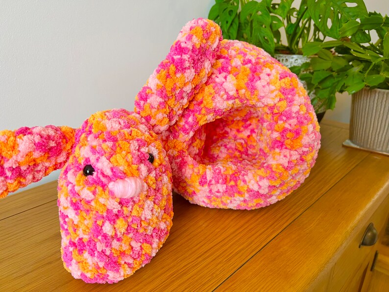 May include: A pink and orange crocheted bunny toy with black eyes and a pink nose. The bunny is lying on its side with its head resting on its arm.