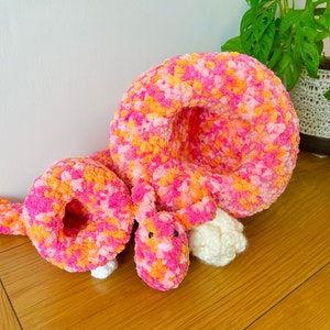 May include: A pink and orange crocheted stuffed animal shaped like a curled up caterpillar. The caterpillar has a white crocheted head and a pink crocheted body.