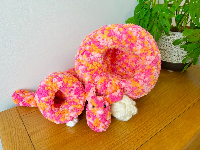 May include: A pink and orange crocheted stuffed animal shaped like a curled up caterpillar. The caterpillar has a white crocheted head and a pink crocheted body.