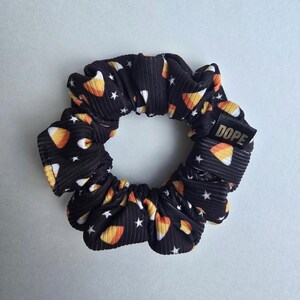 May include: Black scrunchie with a candy corn and star pattern. The scrunchie has a gold label that says "DOPE".