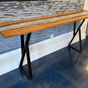 May include: A long, wooden table with a white resin inlay. The table has a black metal X-shaped base.