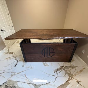 Puede incluir: Un escritorio de madera hecho a medida con una tapa de borde vivo. El escritorio presenta una base de madera marrón oscuro con un marco de metal negro y un monograma. El escritorio está en una habitación con piso de mármol blanco y gris.