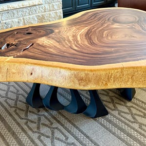 May include: A large, natural wood coffee table with a live edge top and a unique, abstract metal base. The table has a dark brown wood grain with lighter brown accents. The base is made of black metal and has a geometric design.