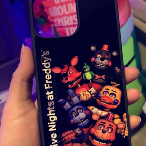 May include: Black phone case with a graphic of characters from the video game "Five Nights at Freddy's" by Scott Cawthon. The characters are Freddy Fazbear, Bonnie the Bunny, Chica the Chicken, Foxy the Pirate Fox, and Golden Freddy.