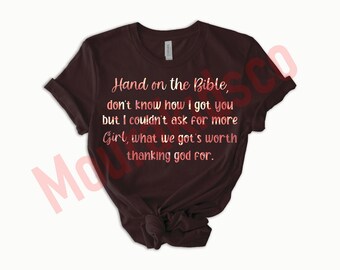 May include: A dark brown short-sleeved t-shirt featuring the text: Hand on the Bible, don't know how I got you but I couldn't ask for more Girl, what we got's worth thanking god for. The lettering is in a light pink and white patterned font.