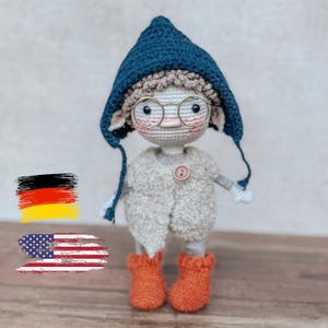 Pode incluir: Boneca de crochê feita à mão usando um chapéu azul, óculos redondos e um casaco branco sujo com um botão laranja. A boneca tem botas laranja e é feita de fio macio. Um item único feito à mão.
