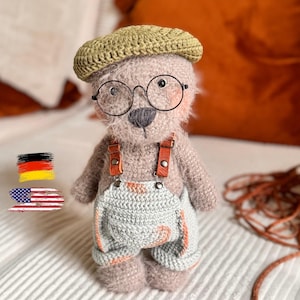 Op de afbeelding: Een pluizige, lichtbruine gehaakte teddybeer met een saliegroene gehaakte baret, een ronde bril met zwart montuur en een lichtgrijze gehaakte tuinbroek met bruine leren bandjes en zilverkleurige metalen knopen.