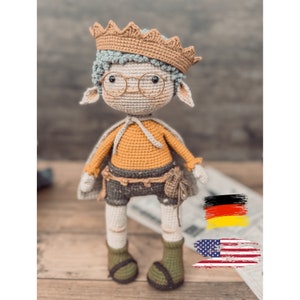 May include: Crocheted doll wearing a crown, glasses, and a cape. The doll has a yellow shirt, gray shorts, and green boots. The doll is standing on a wooden surface.